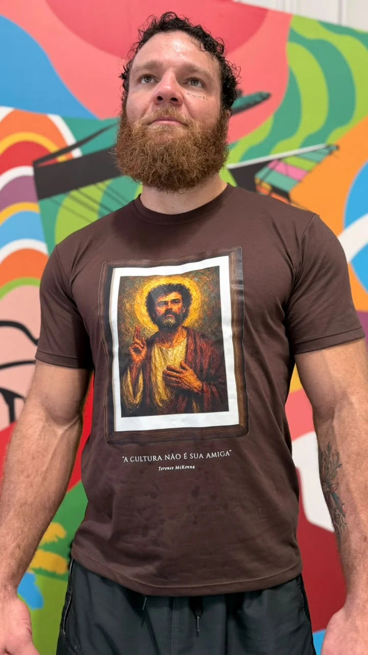 Camiseta marrom Santo Psicodélico no corpo, com estampa de arte em moldura, fotografada em frente a um mural colorido.